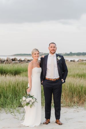 A beach wedding couple stands together on sandy shores with marsh grass and water in the background.