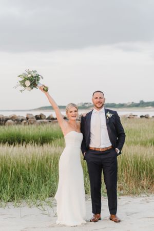Happy newlyweds celebrate their wedding day on a scenic beach with tall grass and ocean views at dusk.