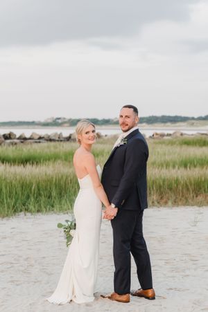 A wedding couple poses together on a beach with coastal vegetation and cloudy skies behind them.
