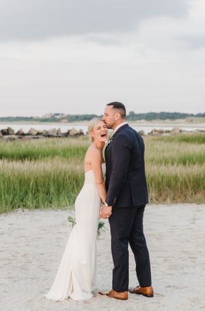 A romantic couple shares a joyful moment on a beach in formal attire with tall grass and cloudy skies in the background.