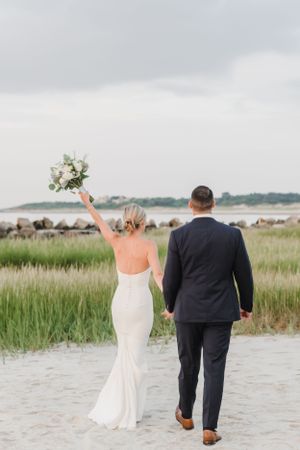 A couple walks on sandy beach path with bride in white gown raising bouquet as grassy marsh stretches behind them.