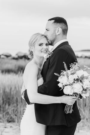 Black and white wedding portrait of newlyweds embracing outdoors with bride holding bouquet of flowers.