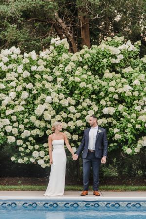 A couple stands by a pool with lush white hydrangea bushes and trees creating a beautiful garden backdrop.