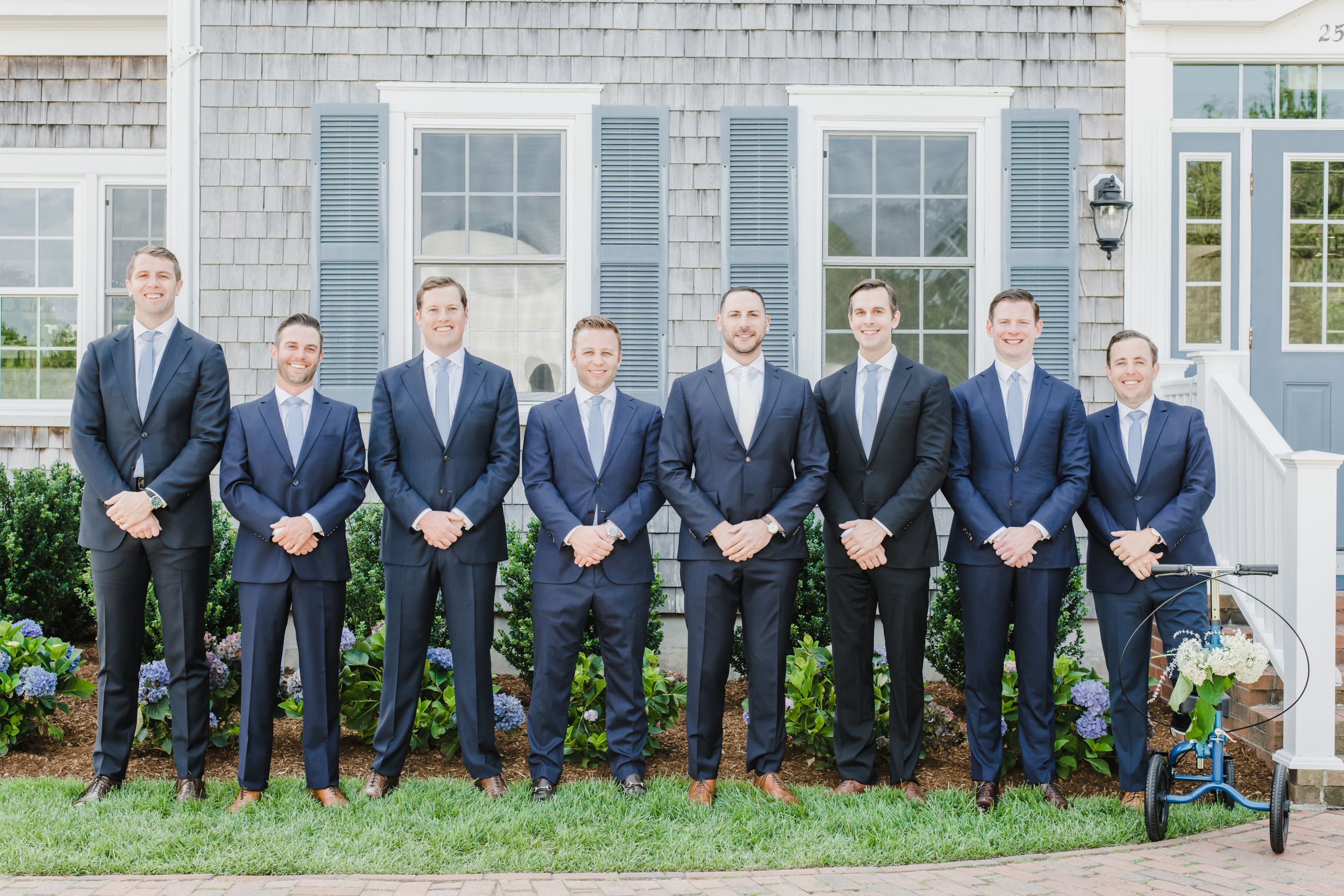 Eight people in matching navy suits and ties stand in a row outside a white brick building with windows.
