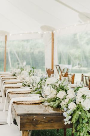 Elegant wedding reception table with rustic wooden farmhouse table, gold chargers, and white floral centerpiece arrangements.