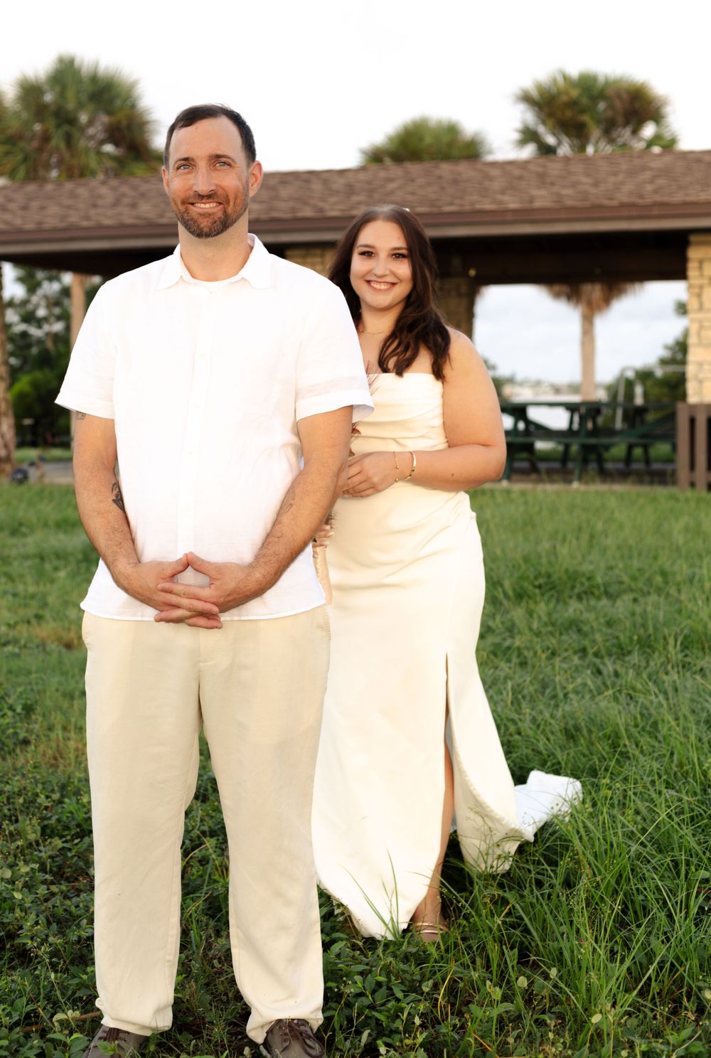 A couple in matching white and cream outfits pose together on grass with a gazebo in the background.