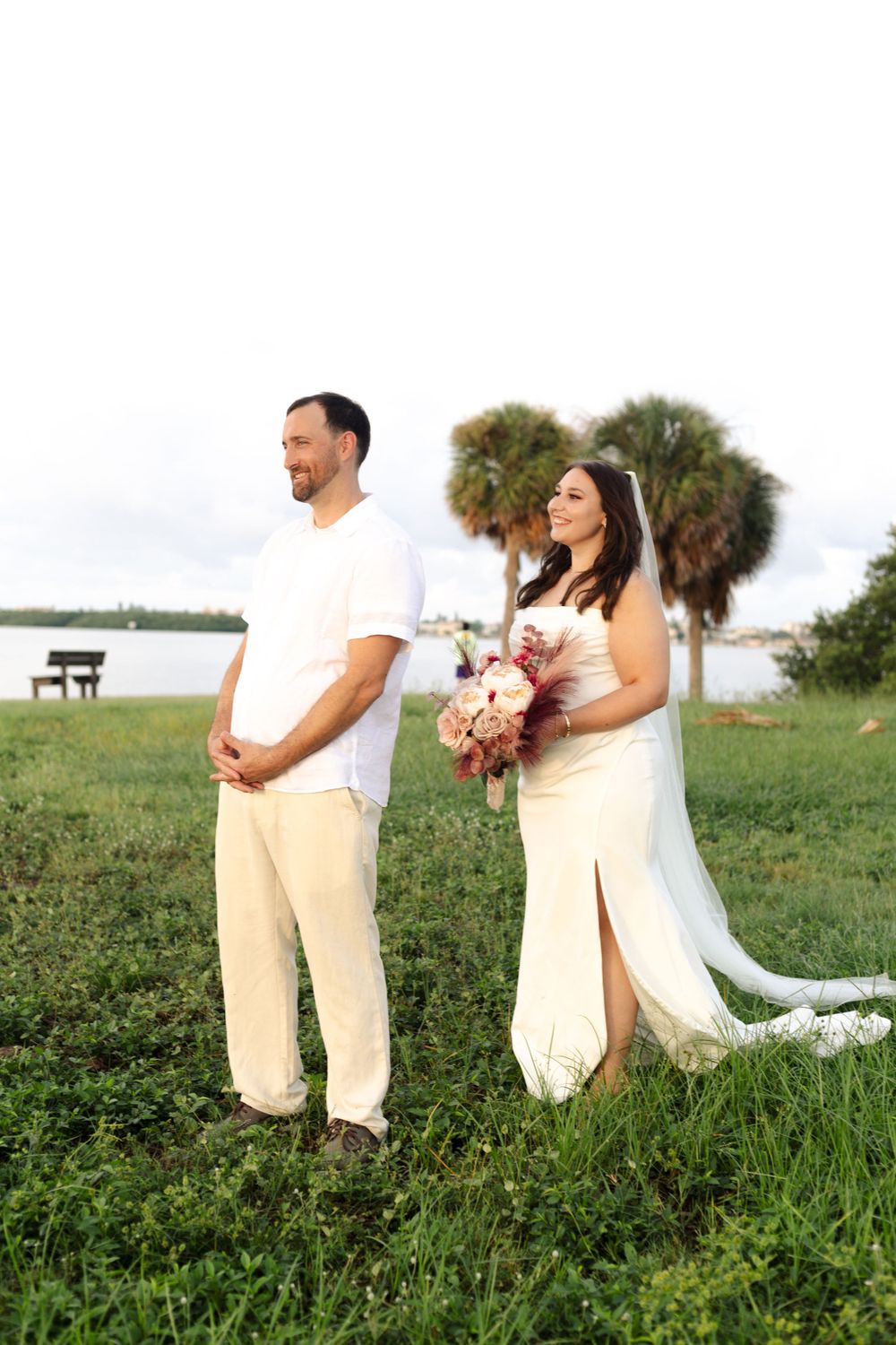 A couple in white wedding attire stands in a grassy field near a waterfront and palm trees at sunset.