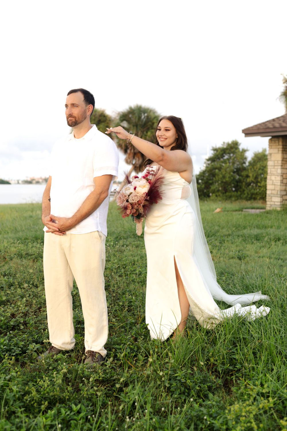 A bride in a white dress playfully poses with someone in matching white attire outdoors on grass during sunset.