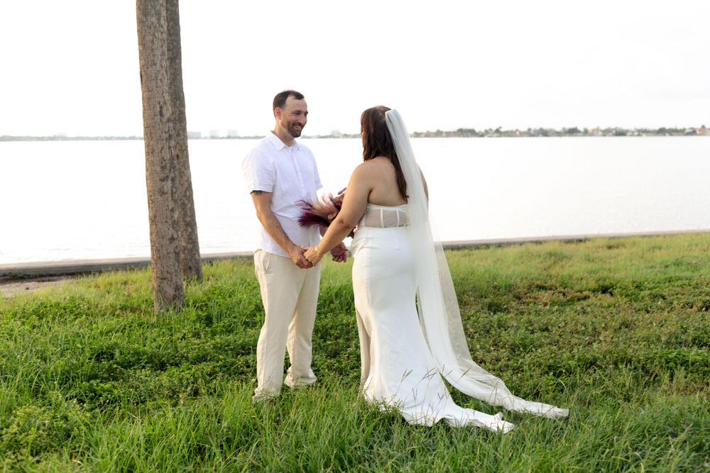 A couple stands on grass near a waterfront during a romantic wedding photoshoot.