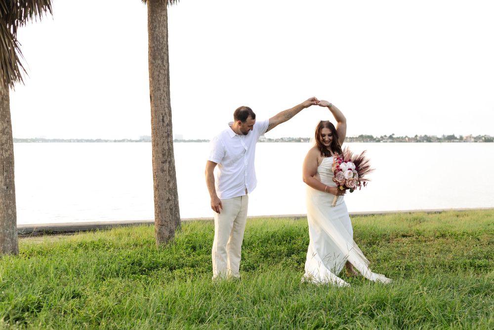 Couple dancing on grassy shore with palm trees and waterfront view during outdoor celebration.