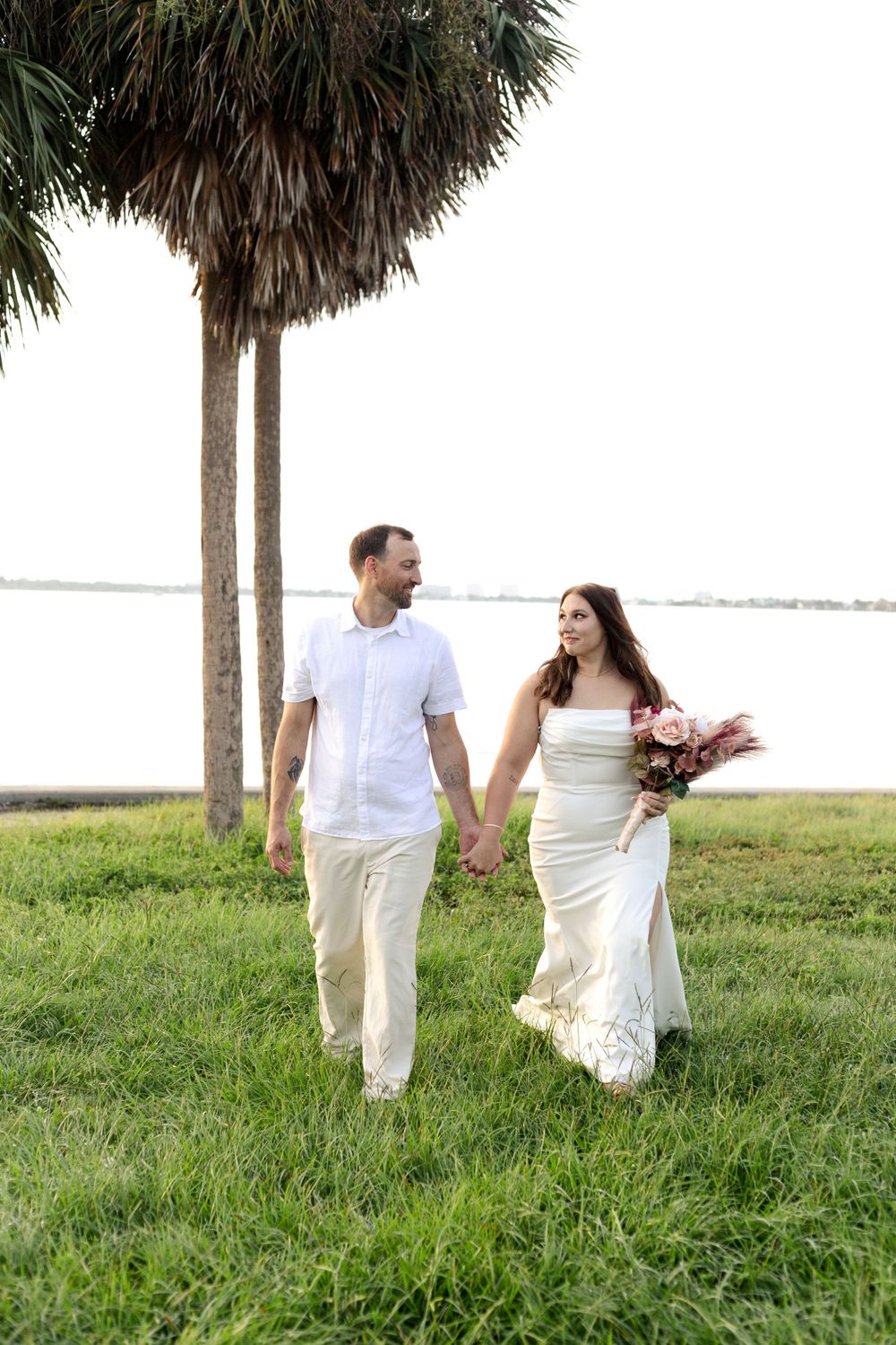 A couple in white wedding attire walk hand in hand on grass beneath palm trees near the water.