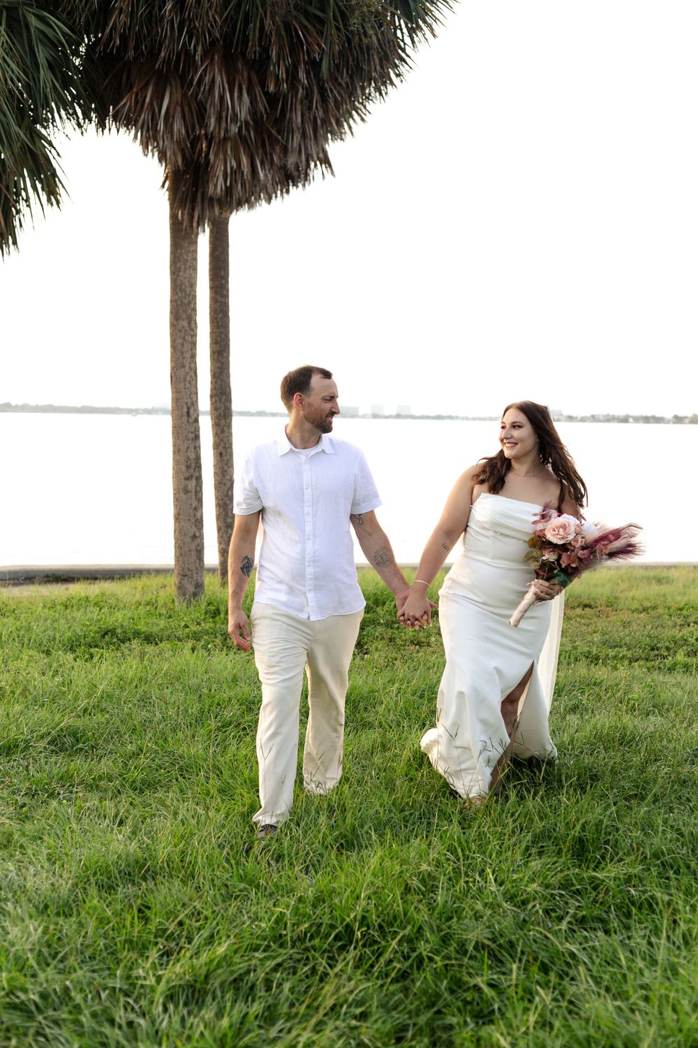 A couple in white attire walks hand in hand on grassy lawn with palm trees and waterfront in background.