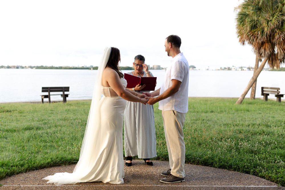 A couple stands with an officiant during a waterfront wedding ceremony at a park with palm trees and benches.