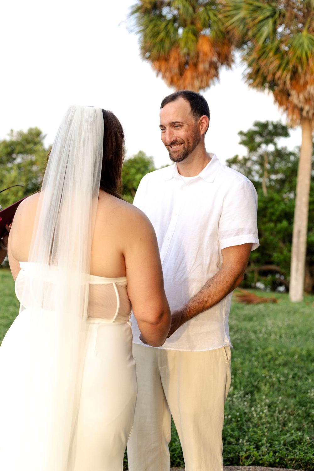 A couple dressed in white exchange loving glances during an outdoor wedding ceremony with palm trees in the background.