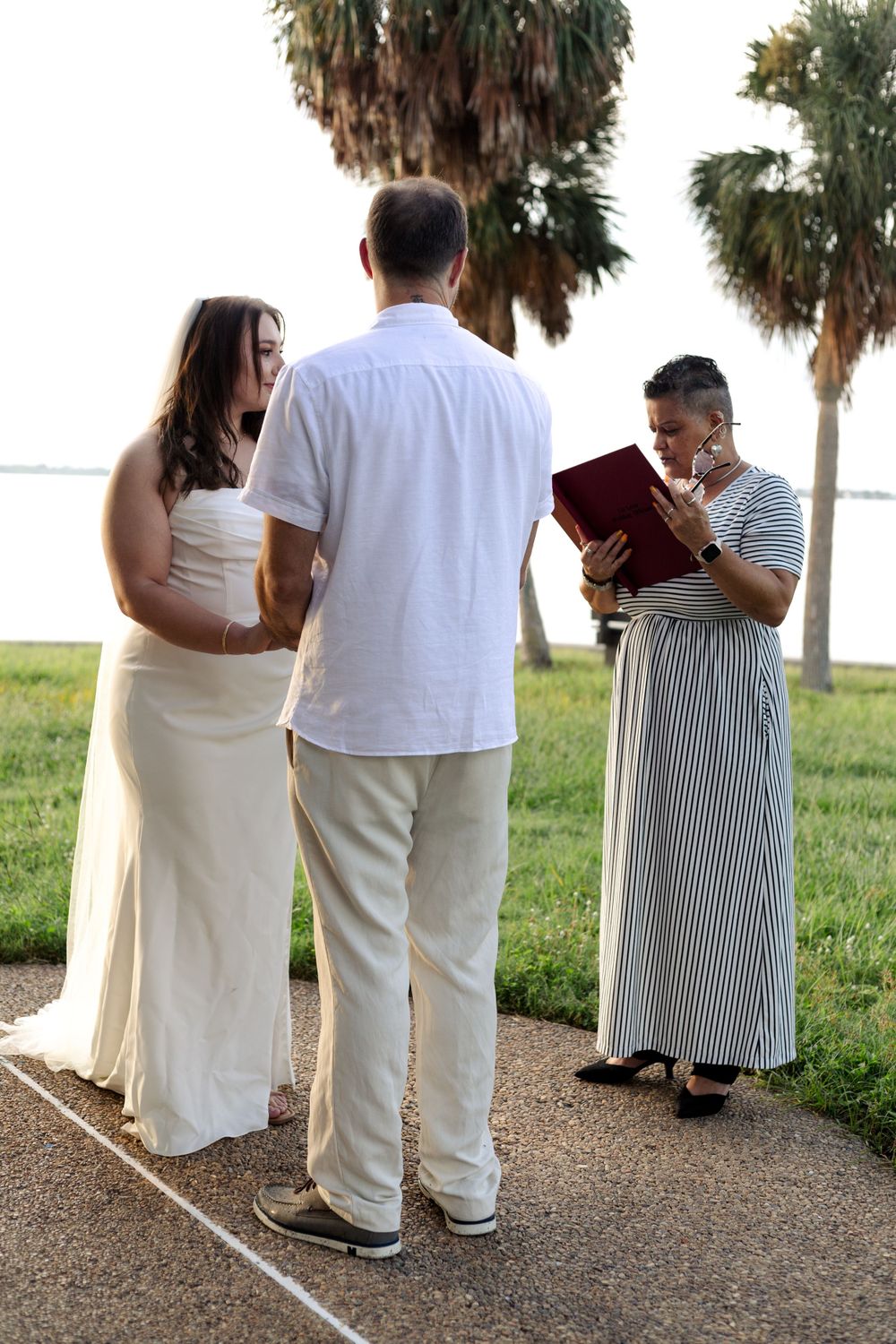 A couple participates in an outdoor wedding ceremony with palm trees in the background on a sunny day.