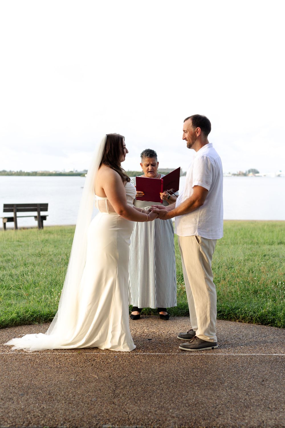 An intimate outdoor wedding ceremony takes place near a lake with officiant and couple on a stone path during sunset.