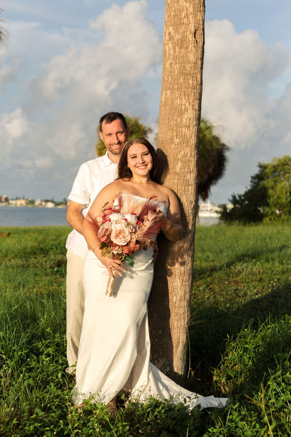 Couple posing for a beach wedding photo at sunset leaning against a palm tree with water in the background.