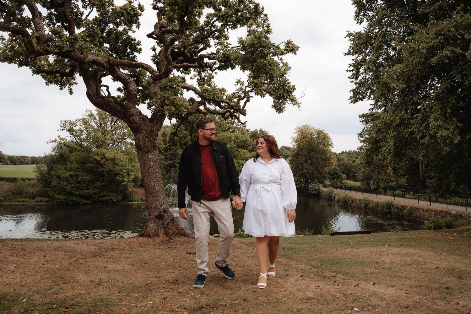 Couple walks hand in hand near a pond surrounded by trees during an outdoor engagement session.