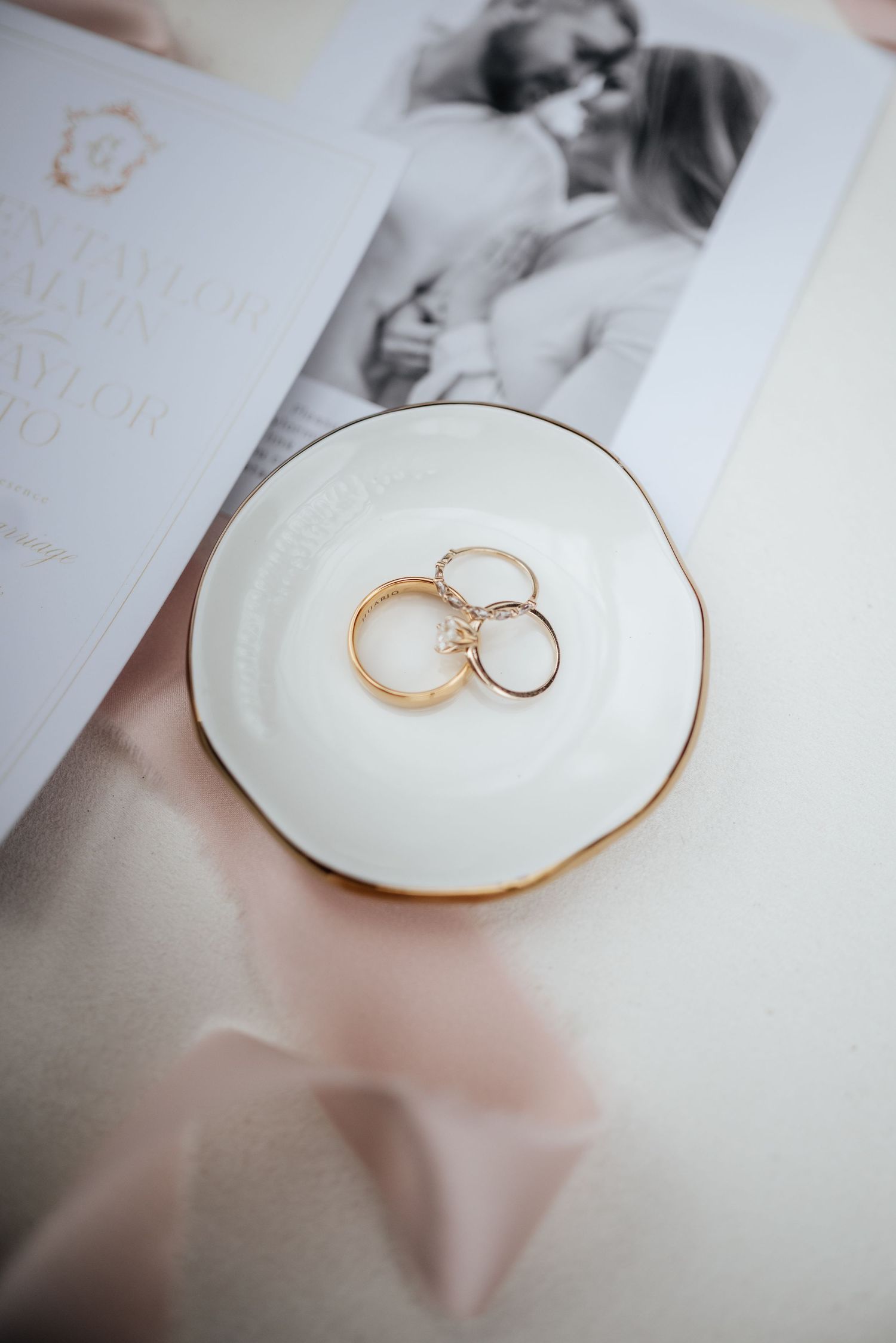 Gold wedding bands and diamond engagement ring displayed on white ceramic dish beside wedding invitation suite.