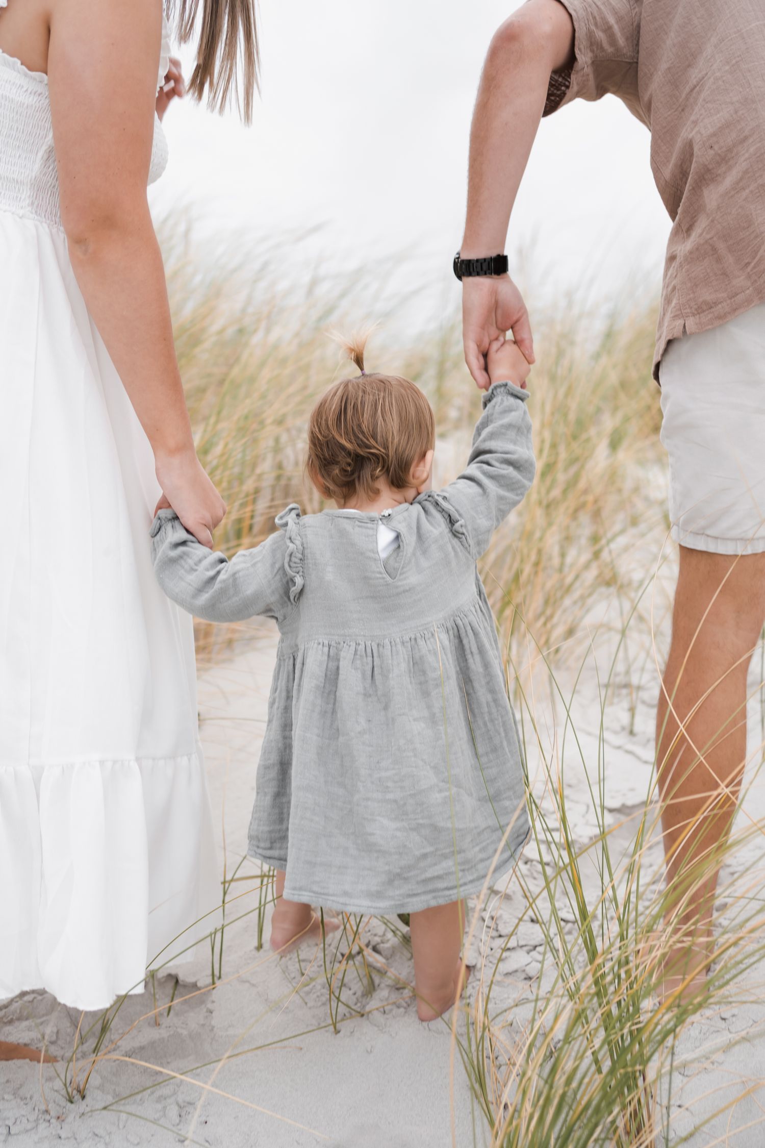 A small child in a grey dress holds hands with adults while walking through beach grass and sand dunes.