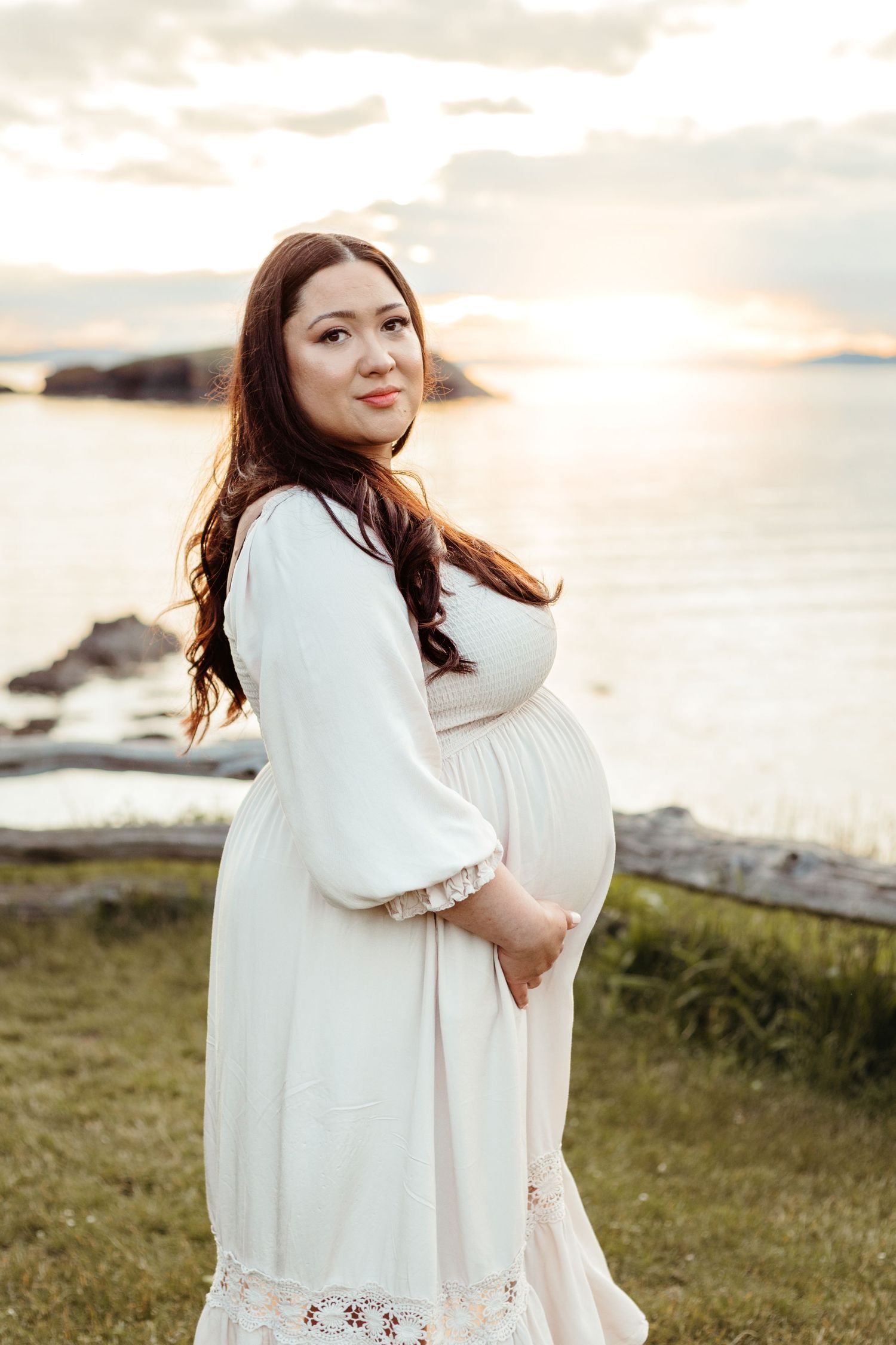 Pregnant woman in white dress poses against sunset coastal backdrop.