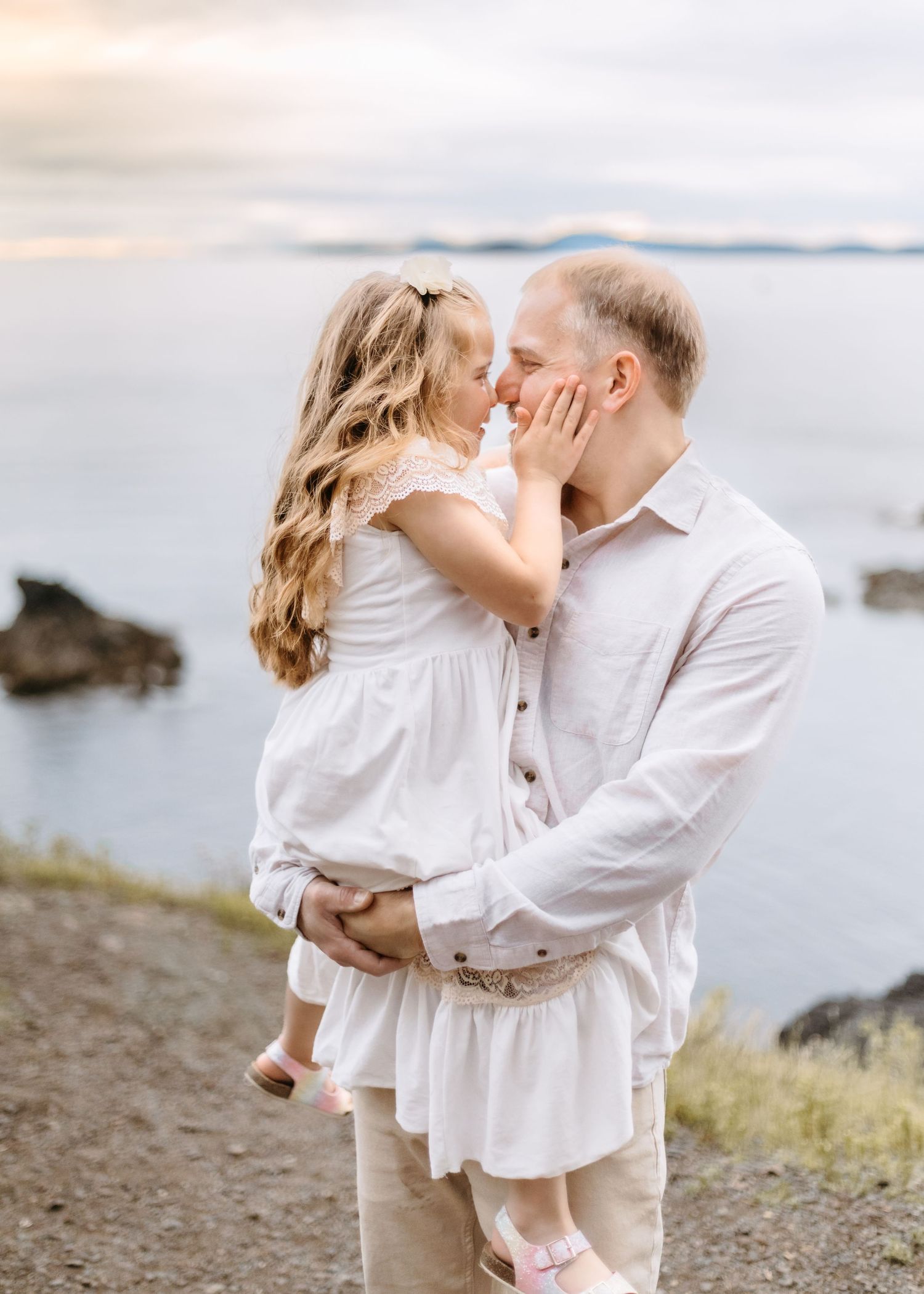 Sweet moment between parent and child in white outfits against coastal backdrop.