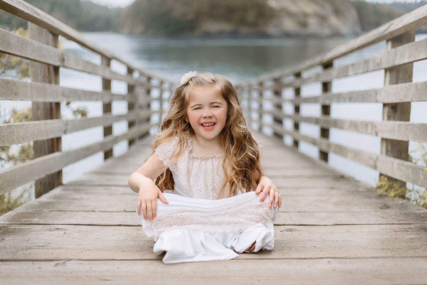 Young child in white dress sitting and smiling on wooden bridge.