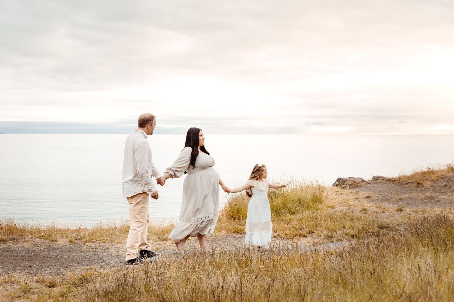 Family of three holding hands while walking along coastal path at sunset.