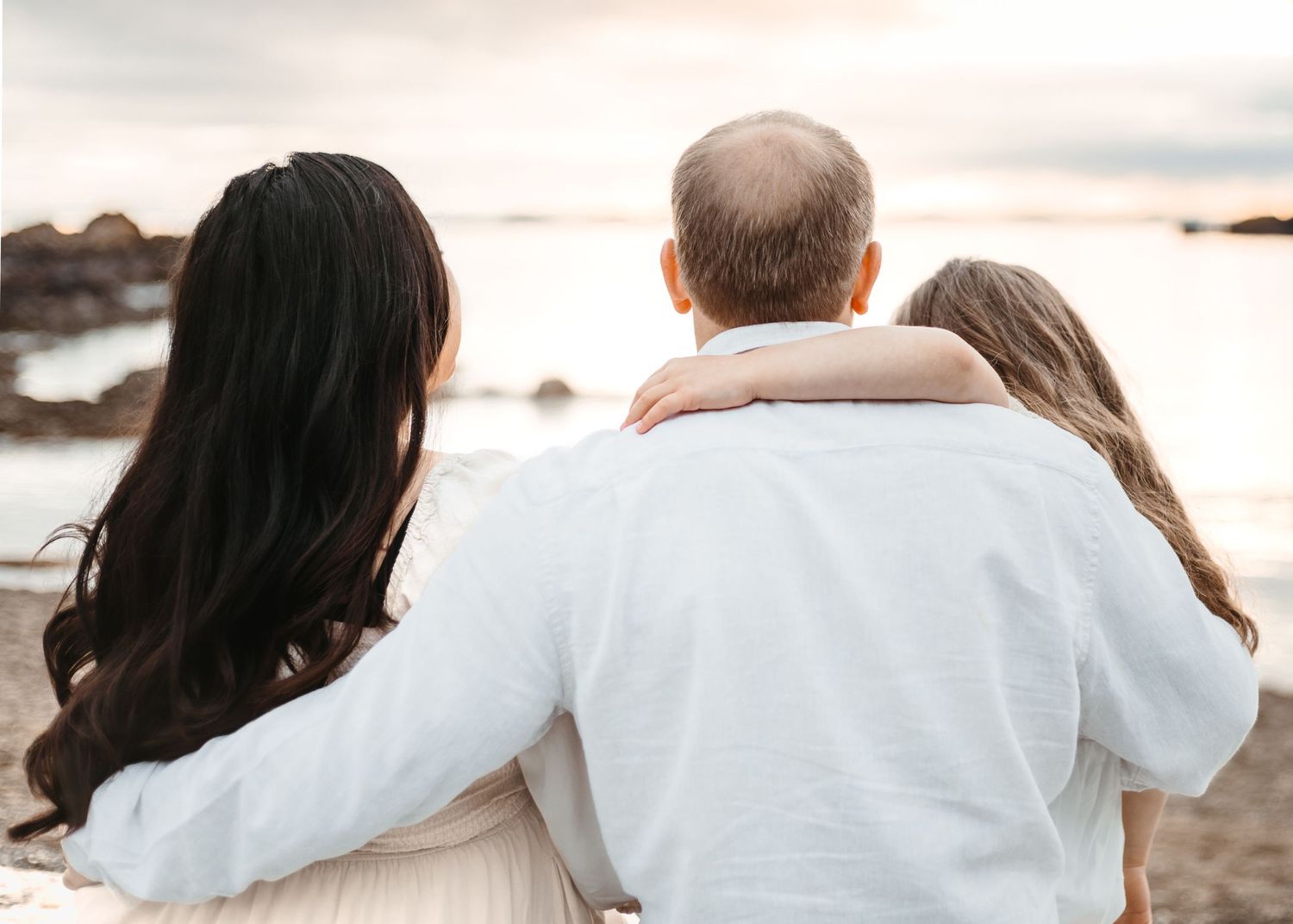 Family enjoying a tender moment together while watching sunset over rocky coastal waters.
