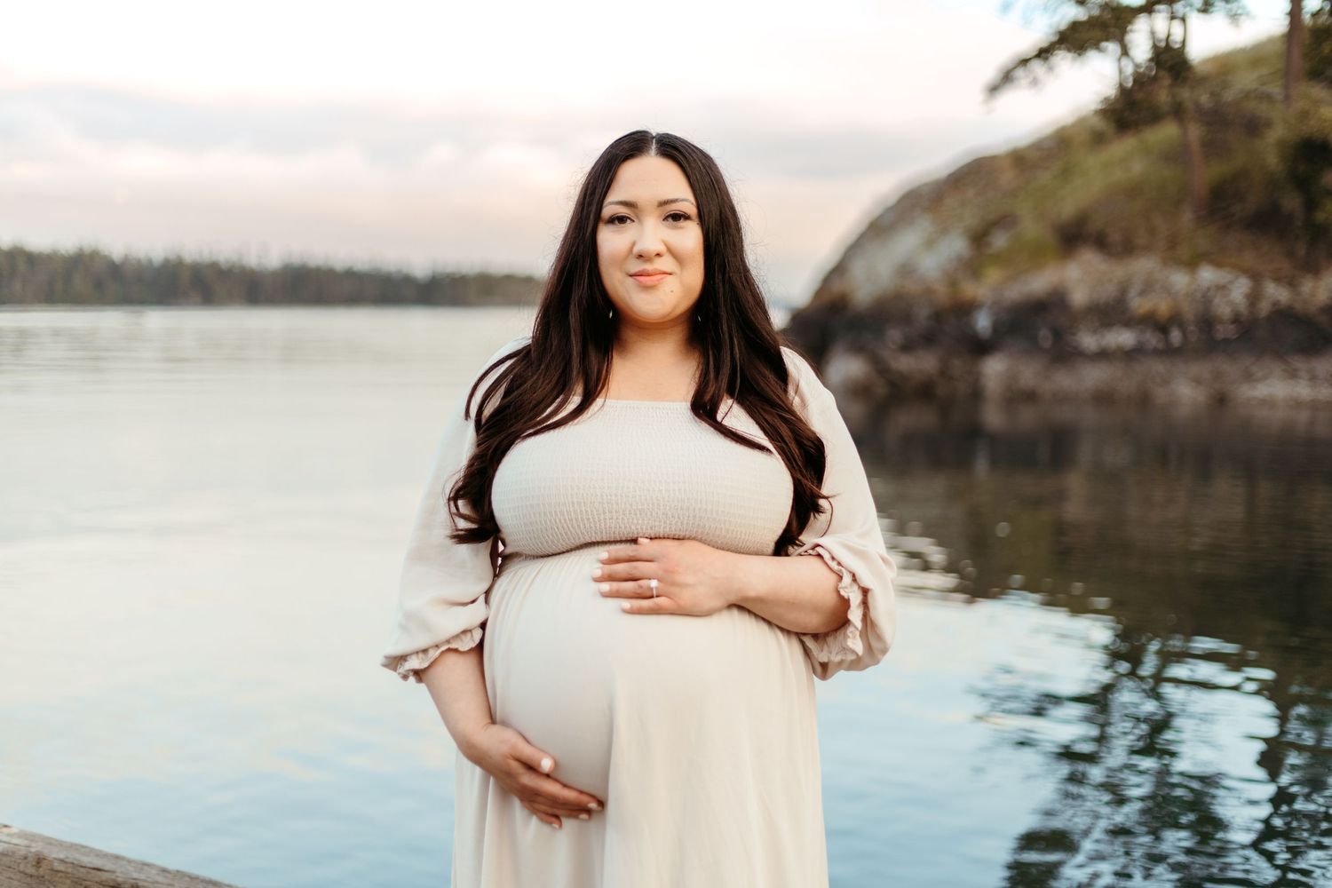 Expectant mother in white dress poses by serene waterfront during golden hour.