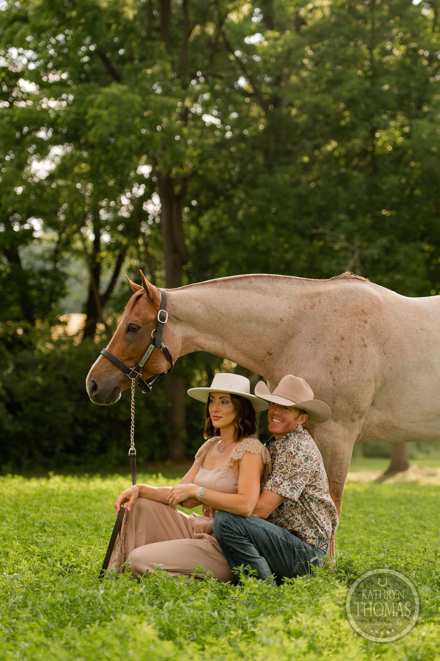 A couple sits in front of their cream-colored horse in a peaceful meadow setting.