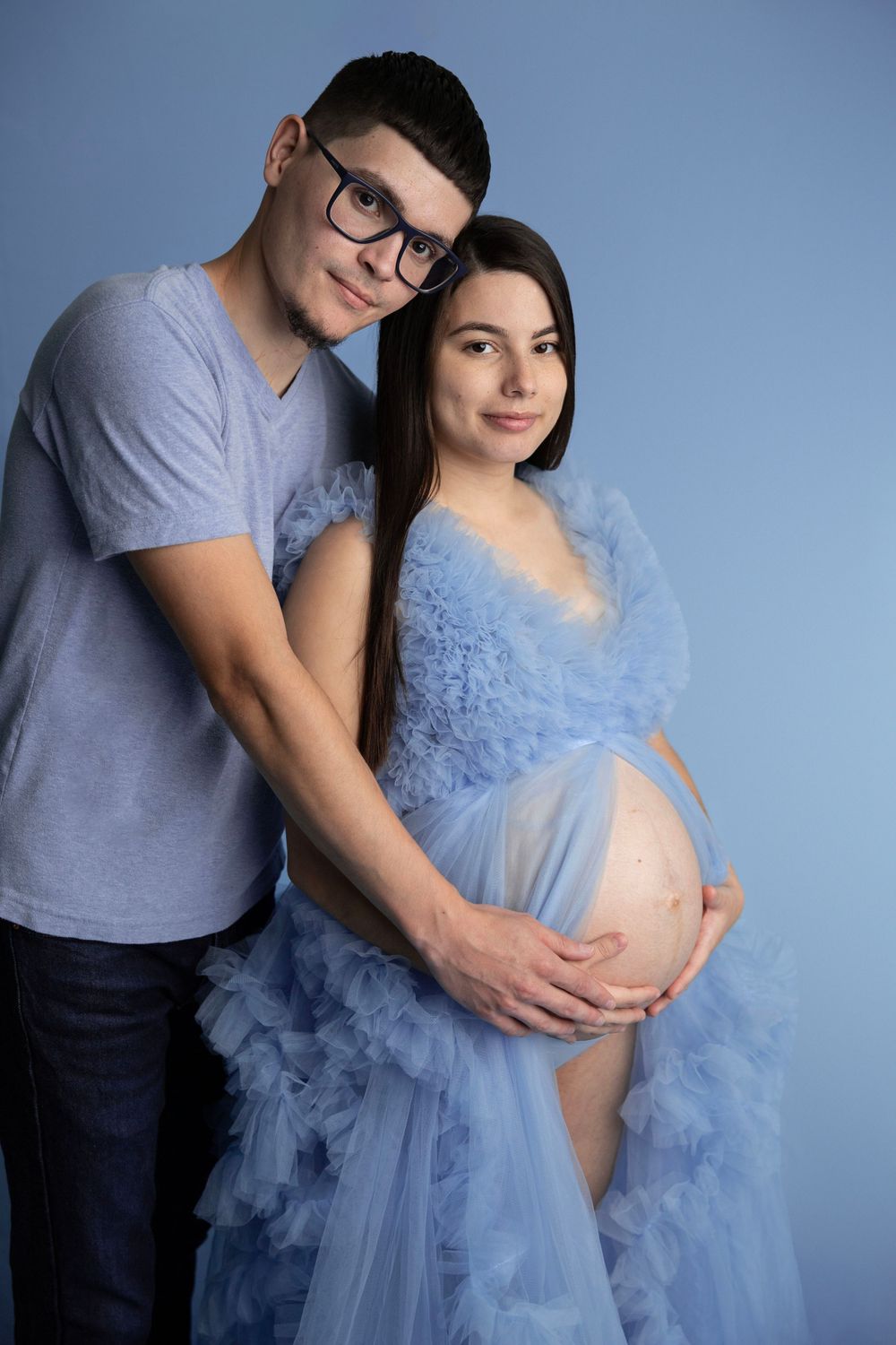 Couple maternity photo with man embracing pregnant woman in blue ruffled tulle gown on blue backdrop.