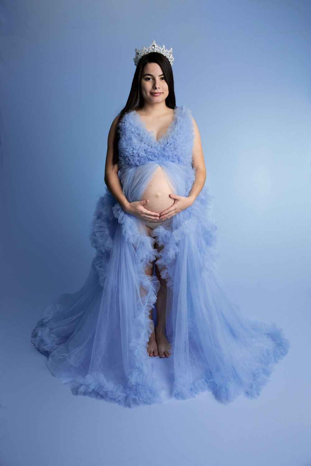 Pregnant woman in blue tulle gown with tiara holding baby bump in studio.