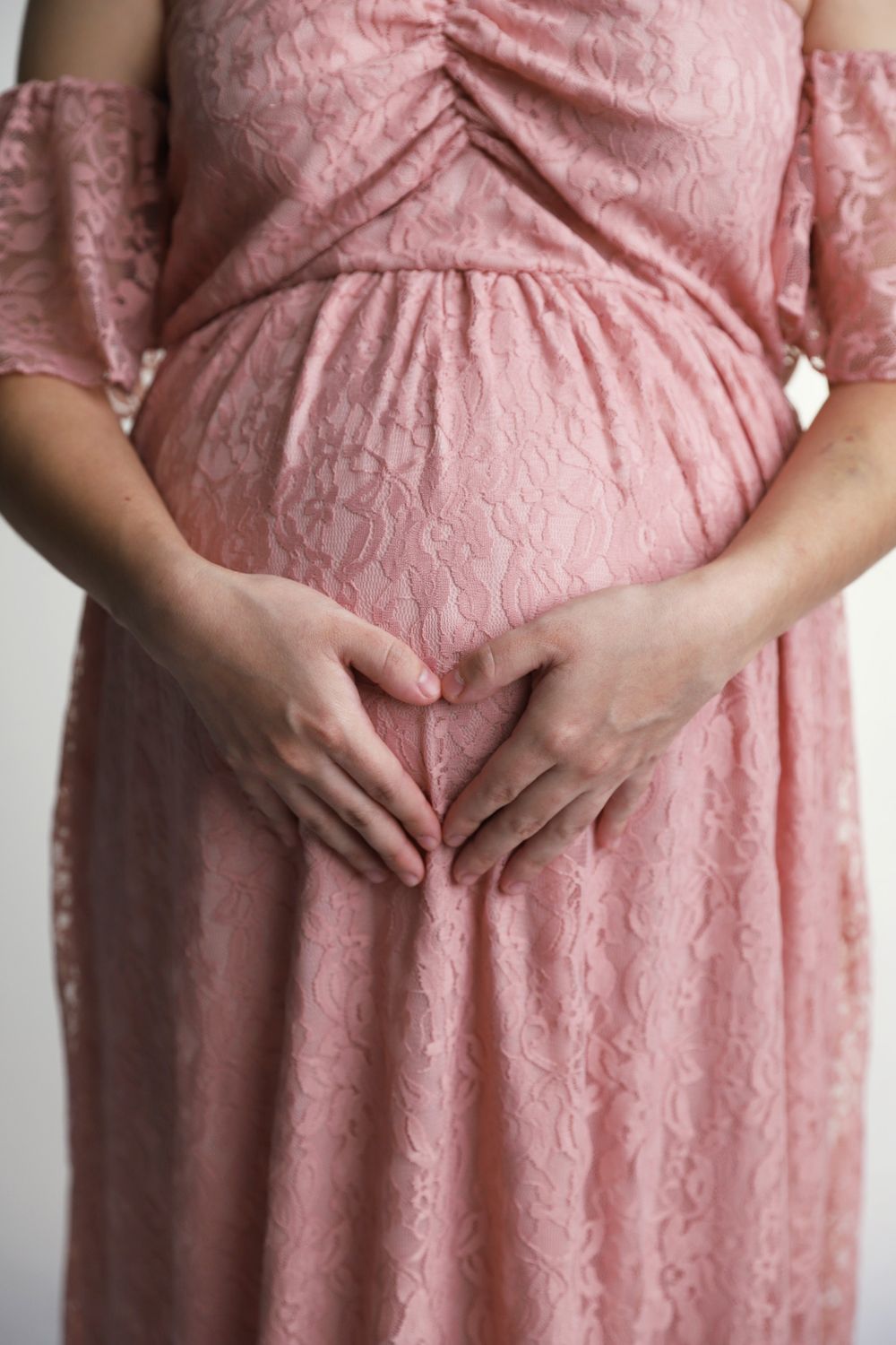 Close-up of pregnant woman making heart shape with hands on baby bump in pink lace dress.