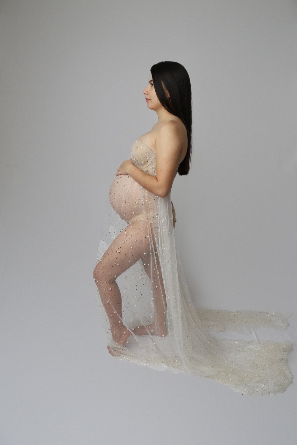 Pregnant woman in sheer pearl-embellished white gown posing in studio maternity shoot.