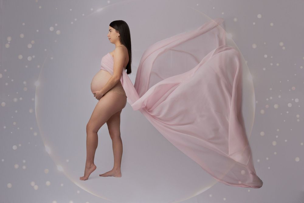 Pregnant woman in pink flowing fabric maternity gown posing gracefully against a soft grey studio backdrop.