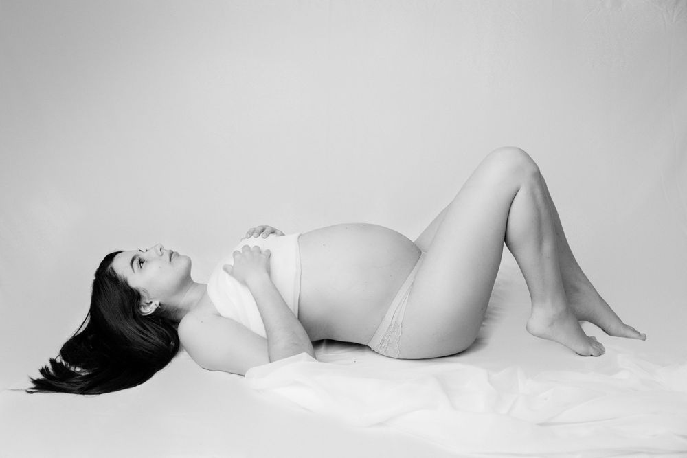 Black and white maternity photo of pregnant woman lying gracefully on white surface.