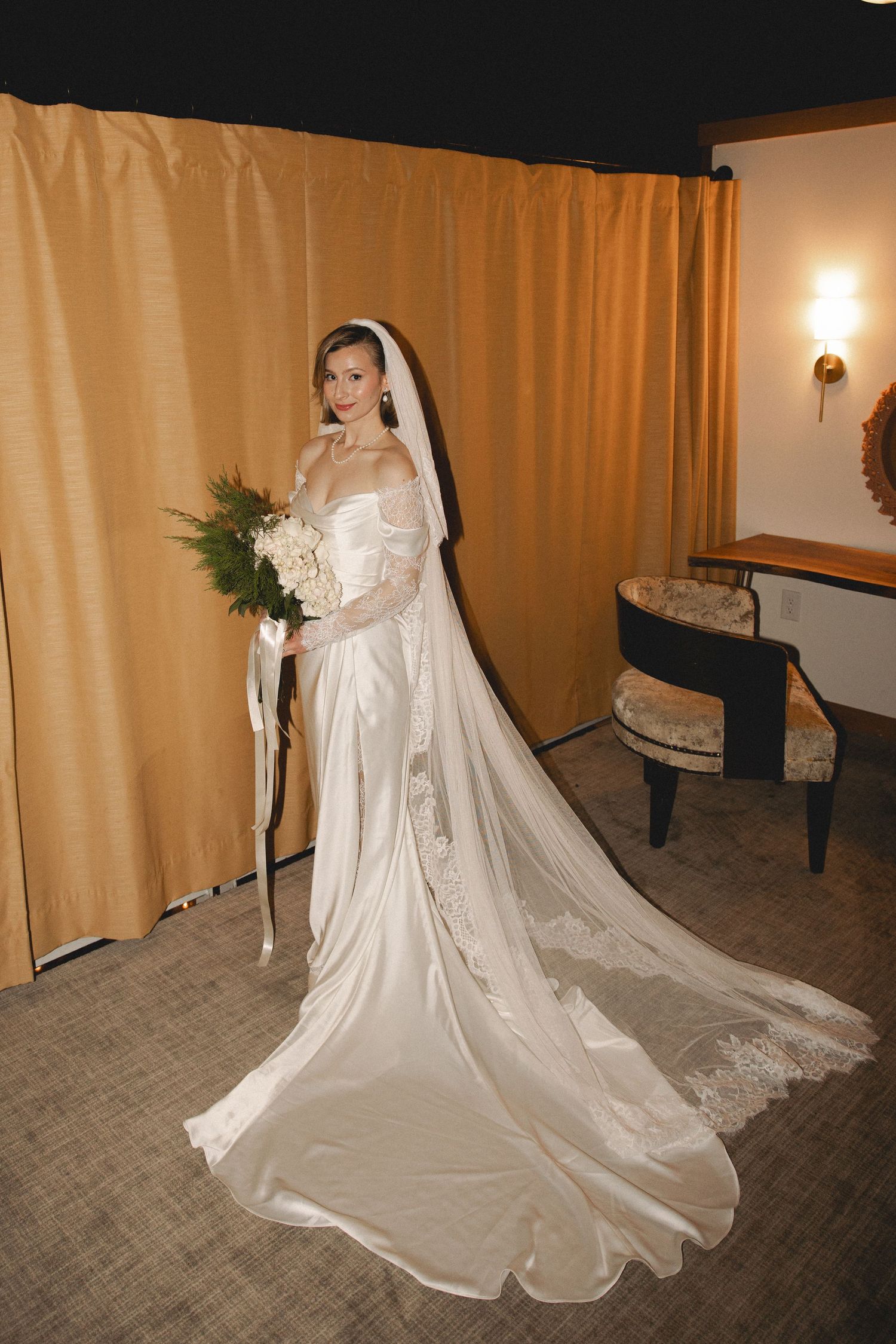 Elegant bride in satin gown with long veil holds white bouquet in a warmly lit room.