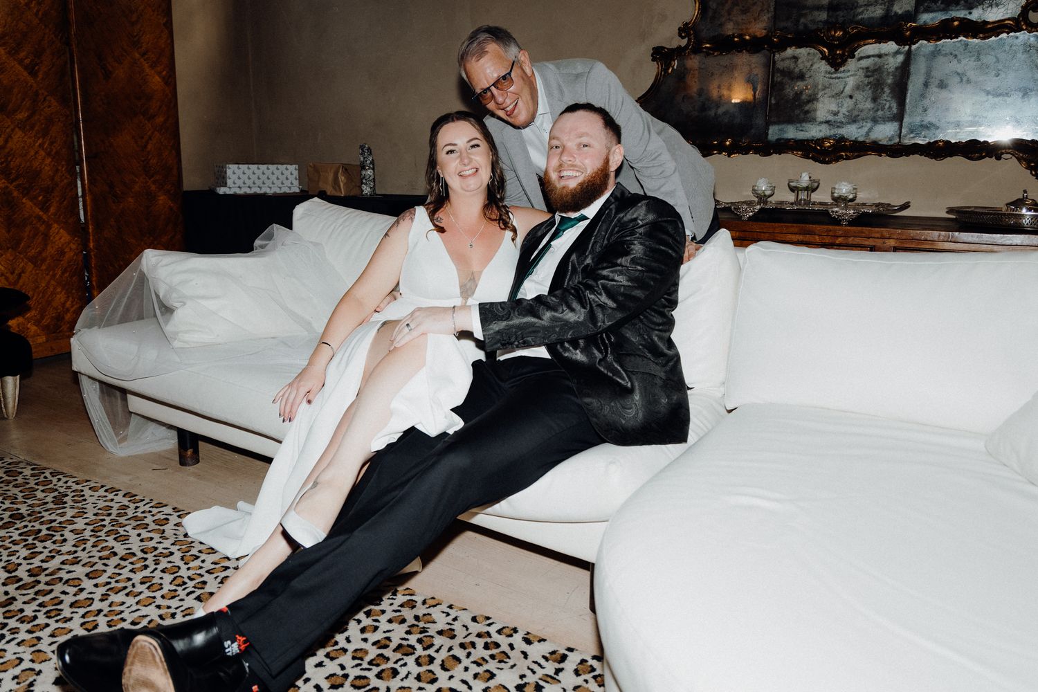 A couple relaxes together on a white couch at an elegant setting, captured by Apertured Media Auckland Wedding Photographer.