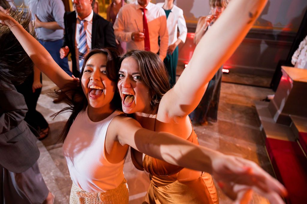 Two party guests dancing energetically together on a dance floor with orange lighting and other people in the background.