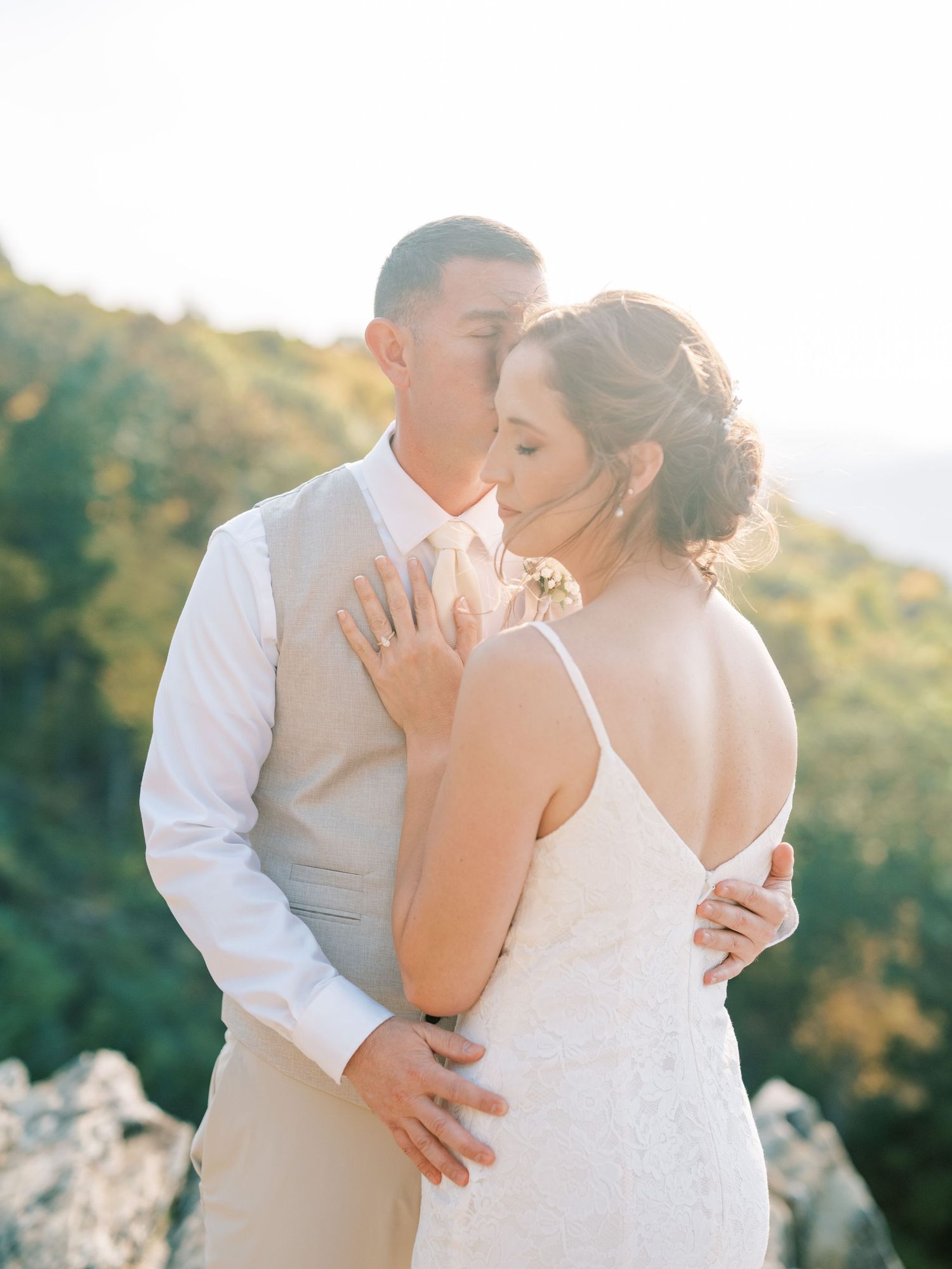 Romantic wedding couple embraces on mountaintop during golden hour as sunlight illuminates natural backdrop.