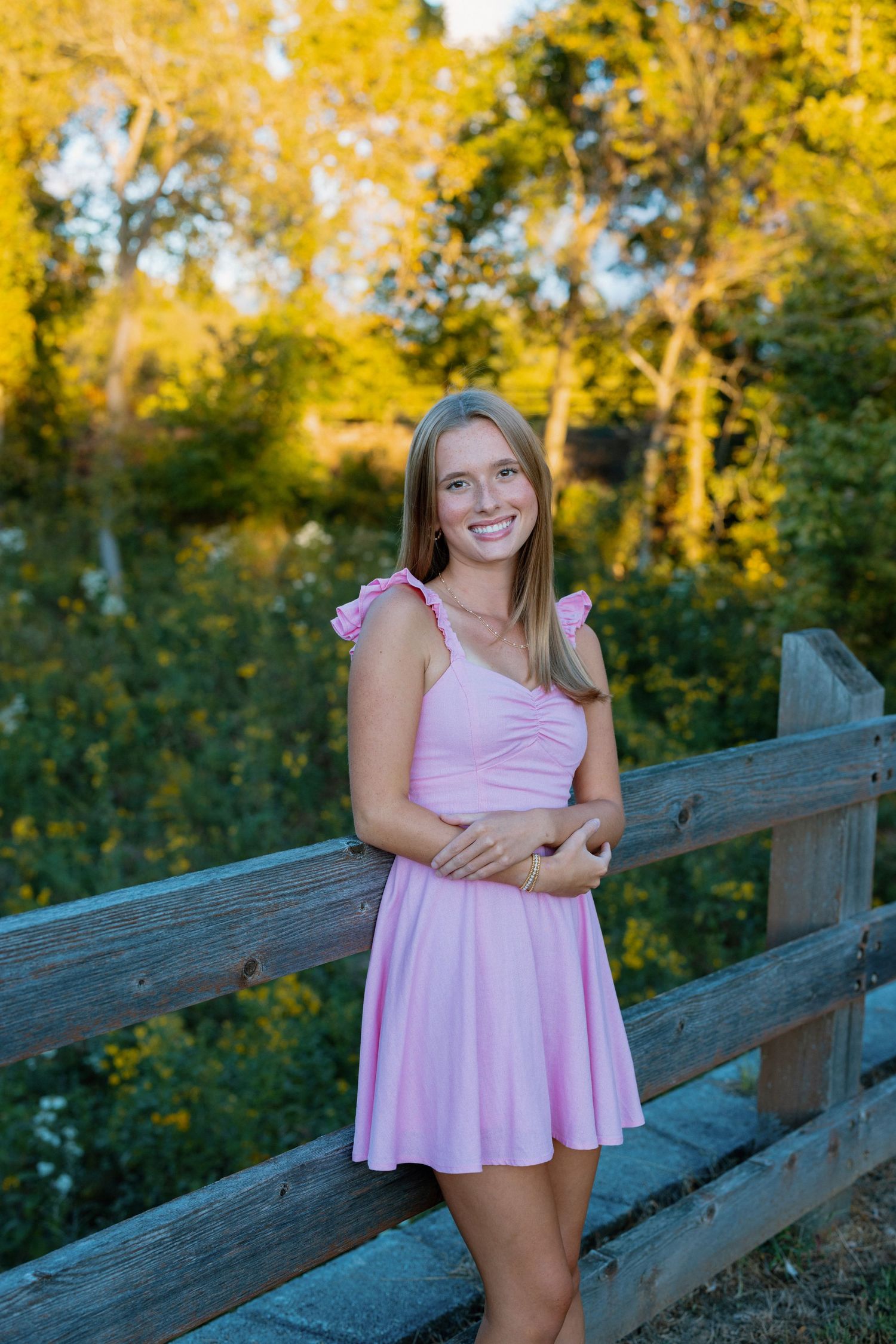 Person in pink dress poses on wooden fence with golden autumn trees in background.