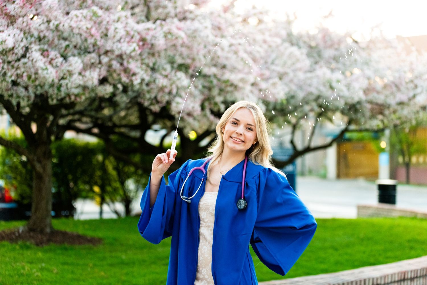 Graduate in blue cap and gown poses under blooming cherry blossom trees in spring.
