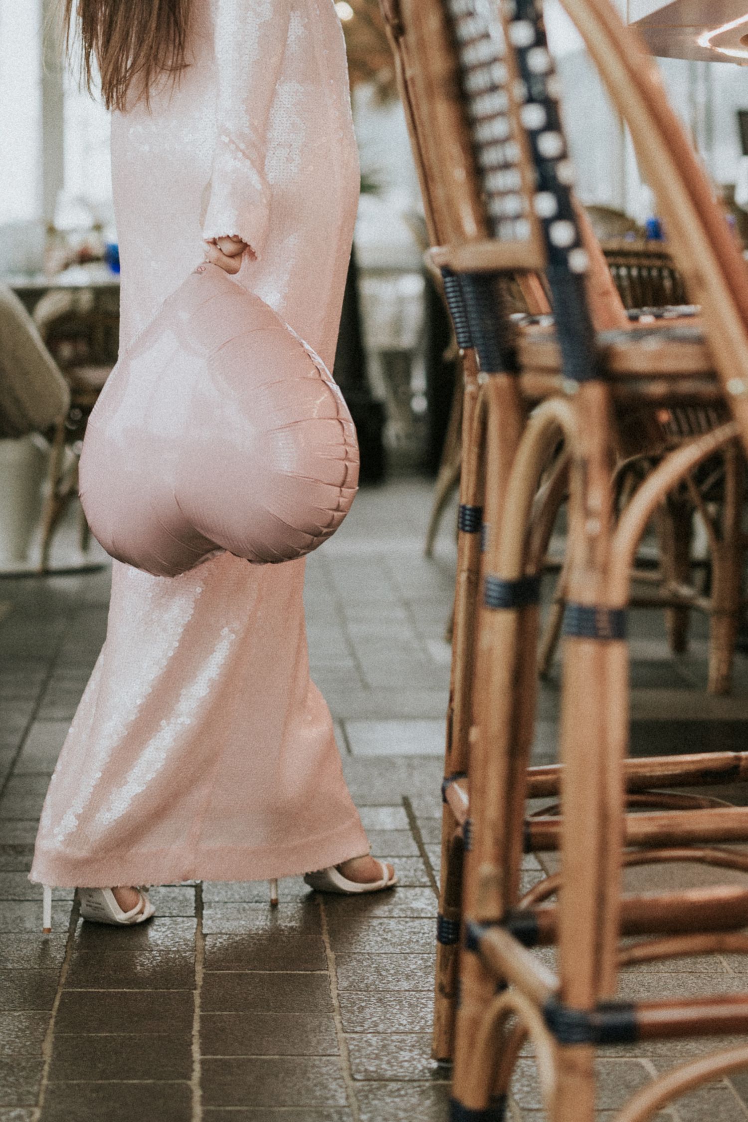 Woman in pink sequin gown holding heart balloon among rattan chairs in elegant restaurant.