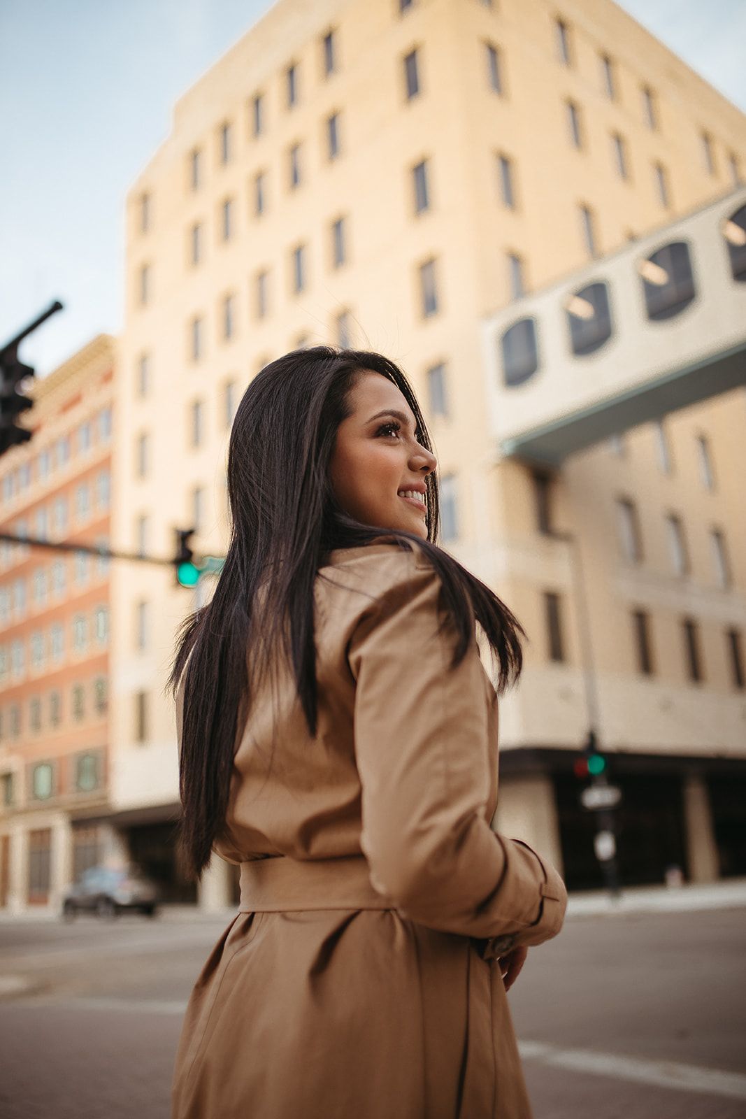 Gal in beige coat smiling while looking over her shoulder on a city street.