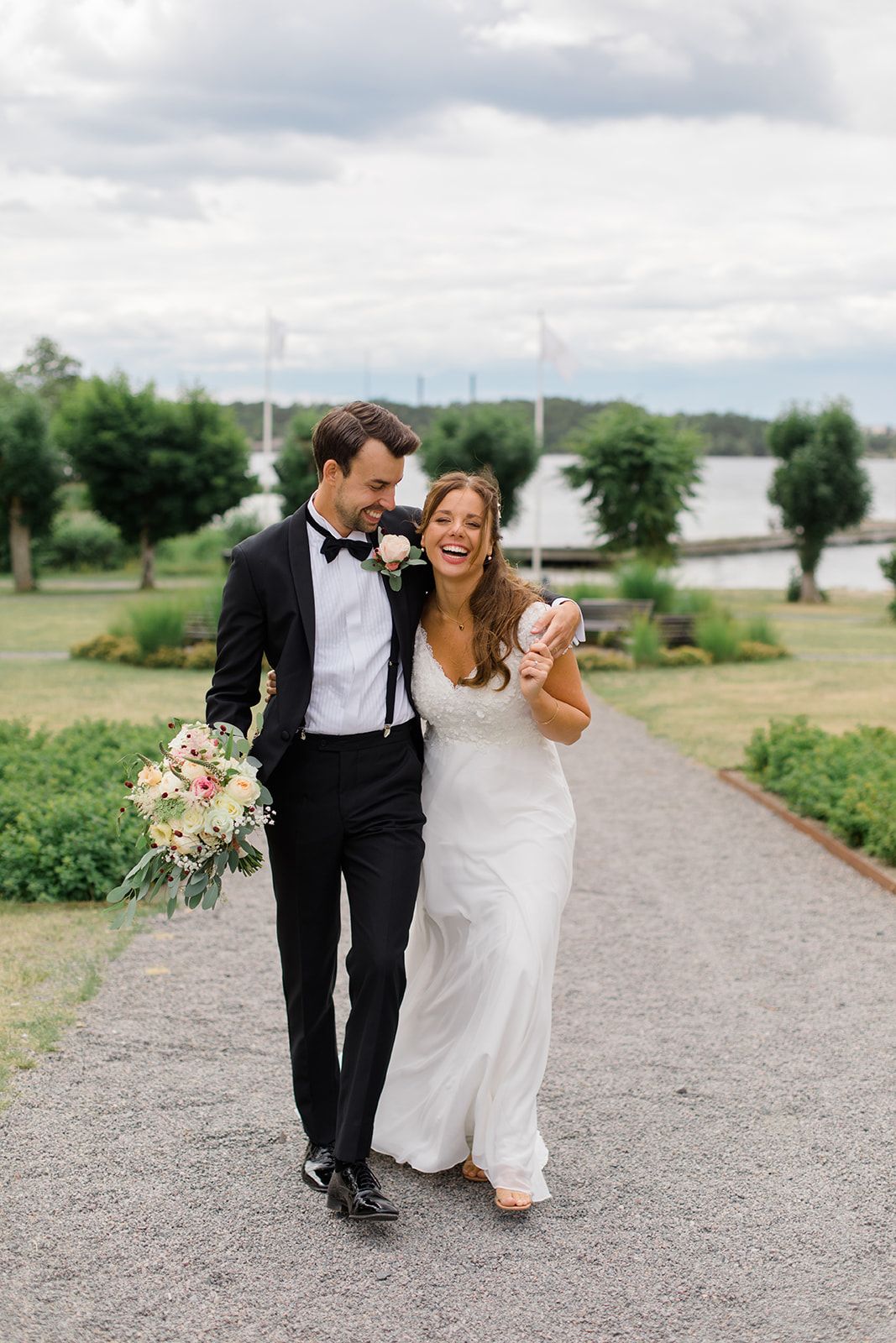 A happy couple walks down a gravel path on their wedding day with boats and trees visible in the background.