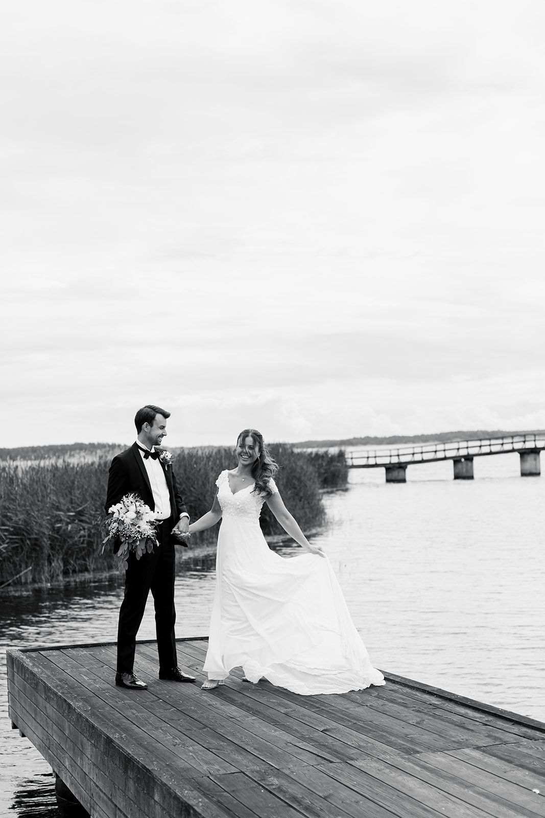 A couple in wedding attire stands on a wooden dock overlooking a lake with a bridge in the distance.