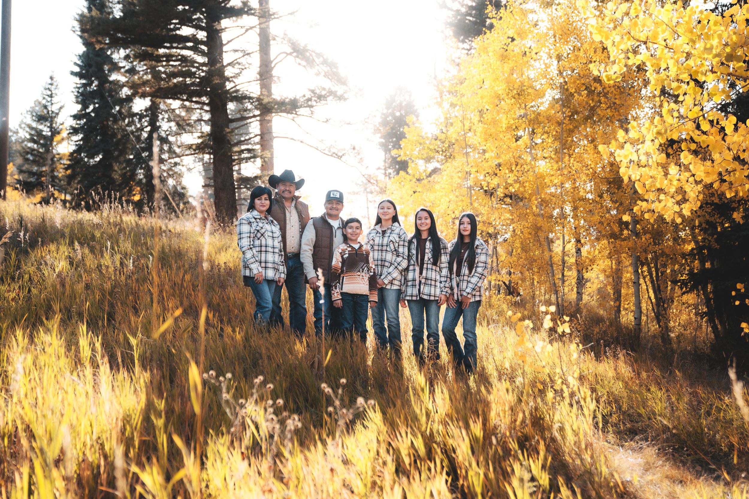 A group of people pose together on a grassy hillside during golden hour with autumn foliage in the background.