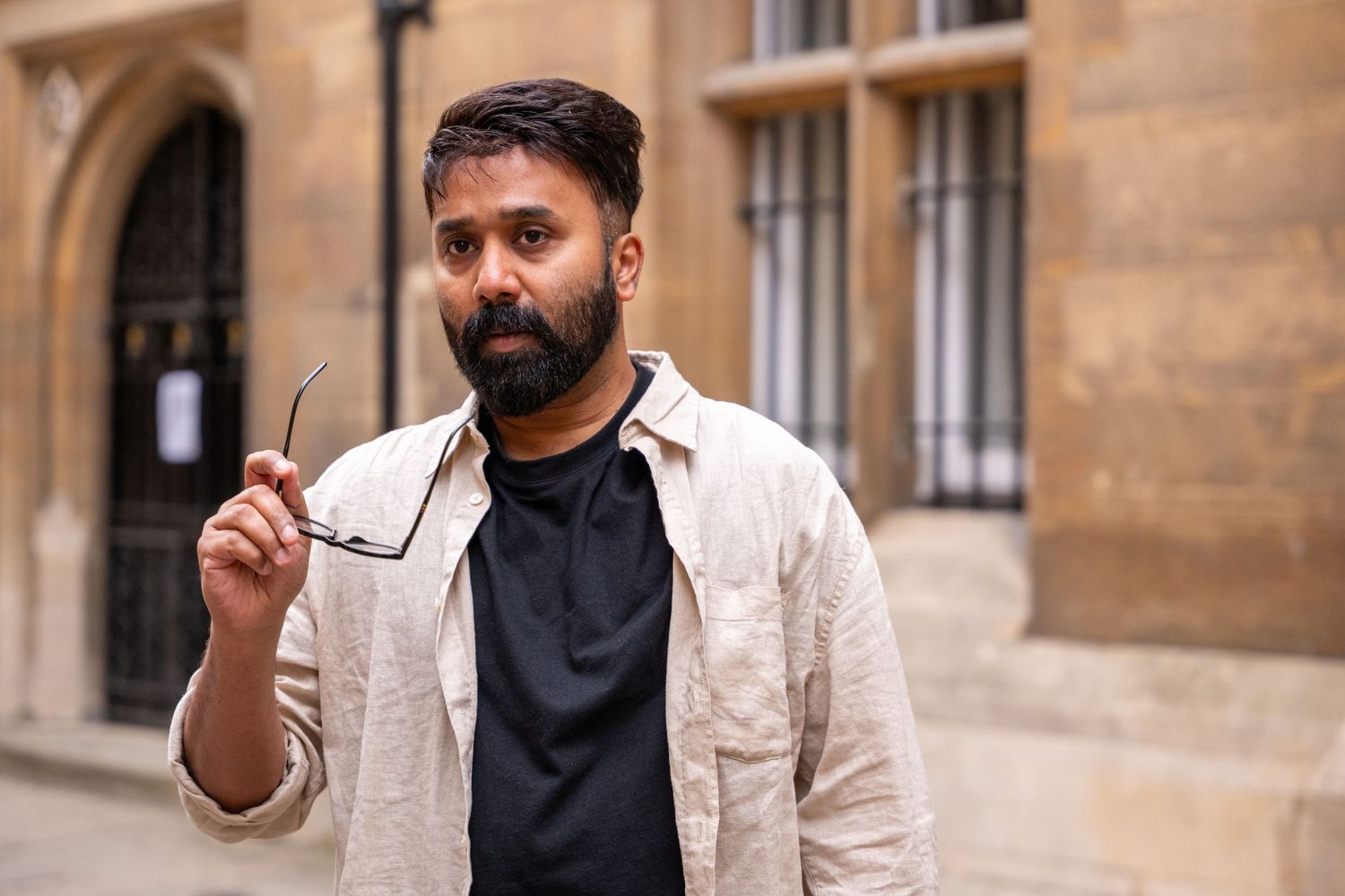A person in a beige blazer and black shirt stands against a sandstone building exterior.