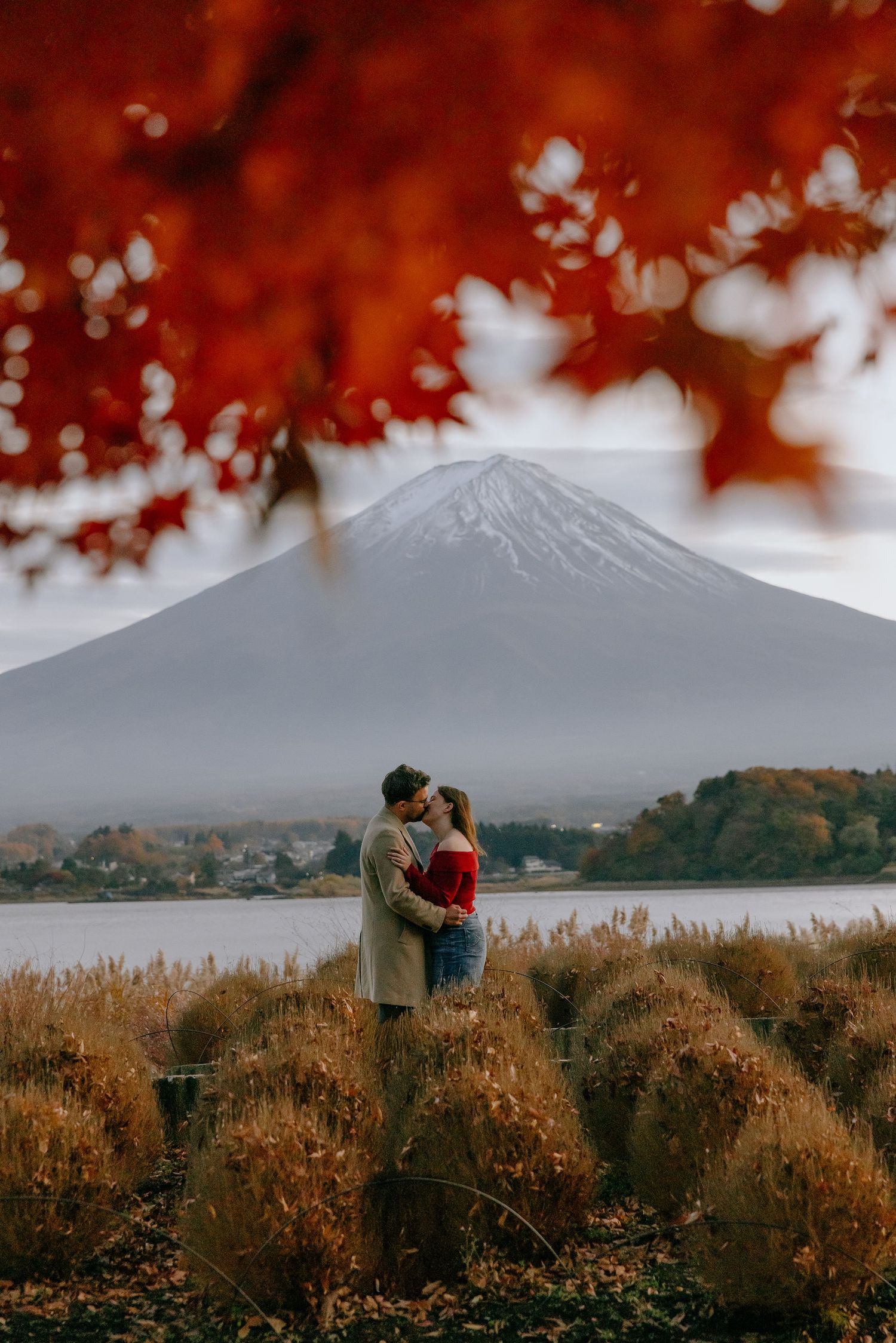 Two people admiring Mount Fuji across a lake with red autumn maple leaves framing the scenic view in Japan.